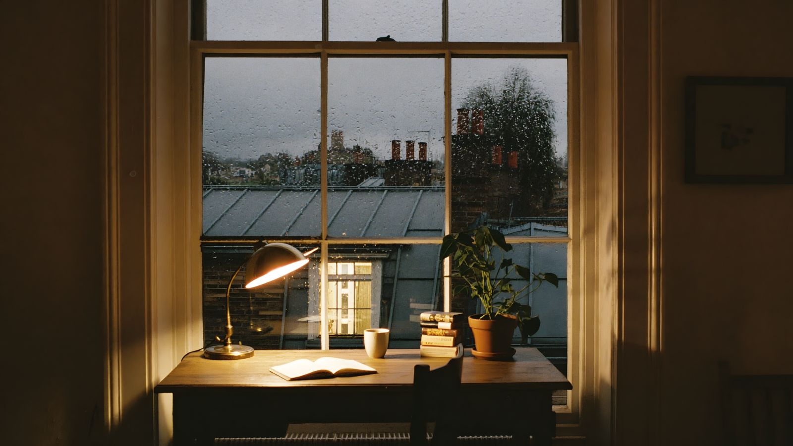 A desk by a rainy window in a London flat, with a lamp on and rooftops outside